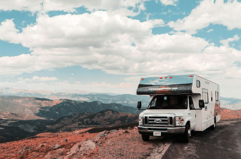 White RV driving along a winding canyon road with mountain formations in the background.