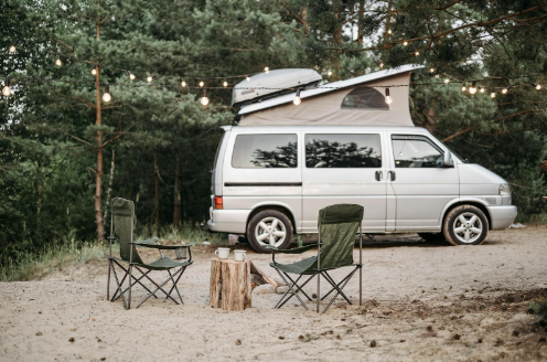 Pop-up camper situated on a camper van next to a campsite with two lawn chairs setup around a wood stump.